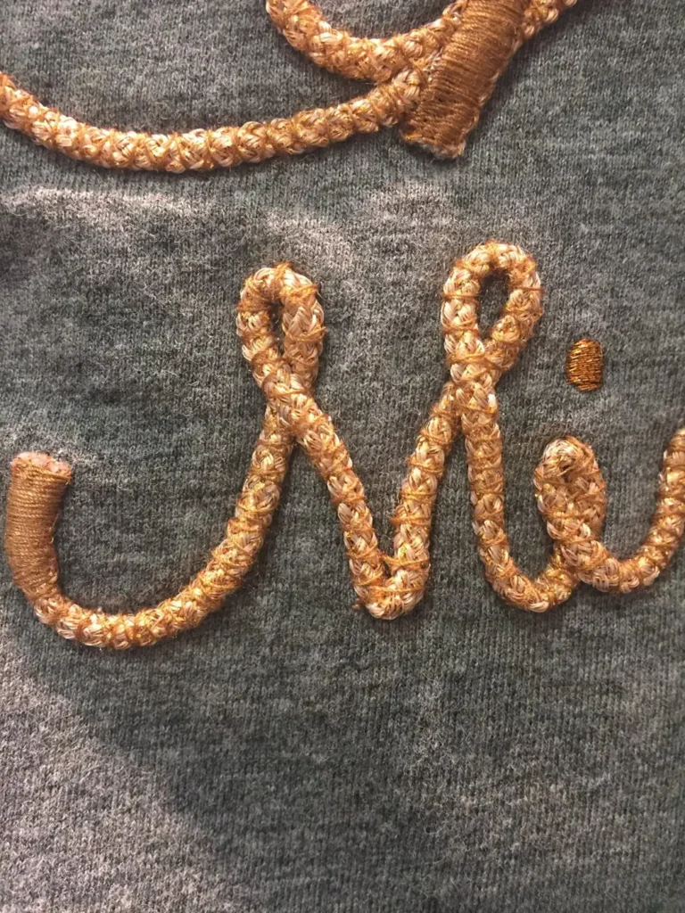 Braided gold cord script lettering on grey knit fabric